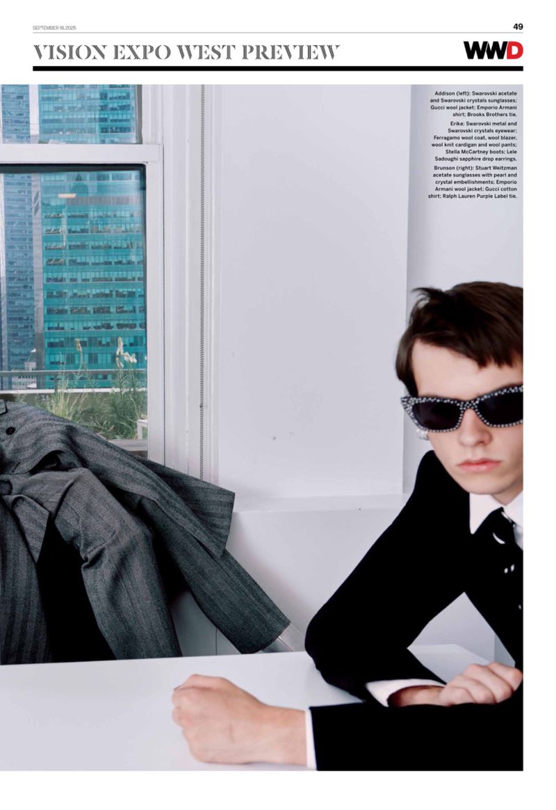 Portrait of a young adult in a dark suit and embellished sunglasses, seated at a white table with a city view through a window.