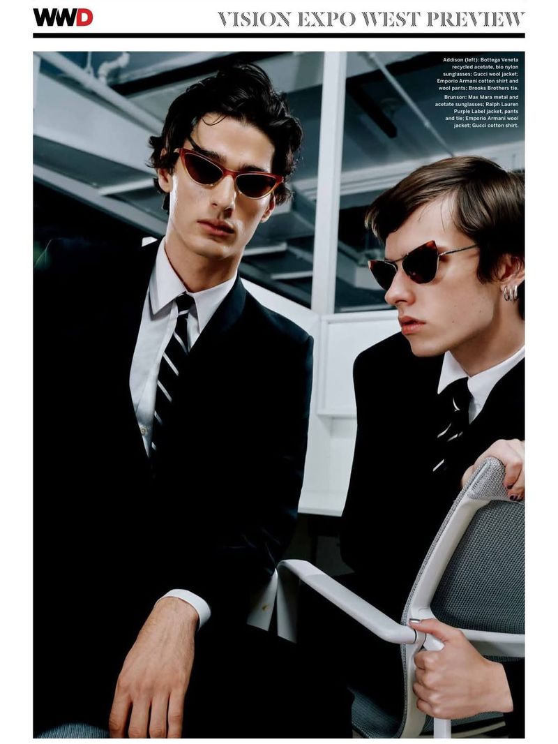Two men pose in suits and sunglasses indoors. One holds a chair. Fashion editorial featuring eyewear and formal attire.