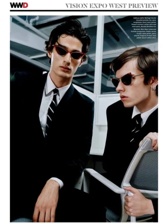 Two young male models in black suits and ties wearing cat-eye sunglasses in an office setting for WWD editorial.