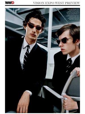 Two young male models in black suits and ties wearing cat-eye sunglasses in an office setting for WWD editorial.