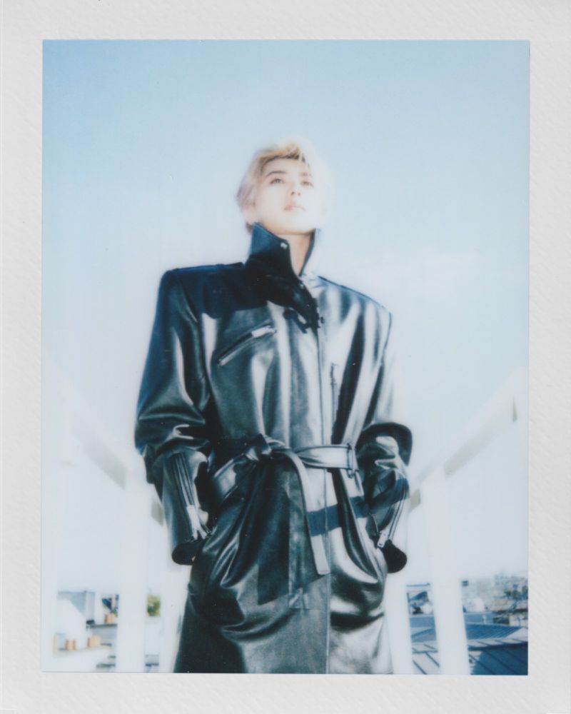 Person in black belted trench coat looking up against a light blue sky, photographed in a polaroid style, wearing gloves.