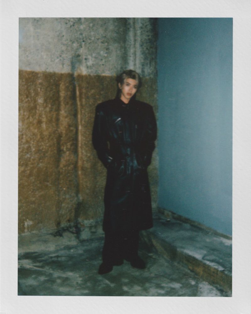 Adult male in black coat poses in dim interior, walls with mixed textures, and a dark ambiance highlighted by a polaroid frame.