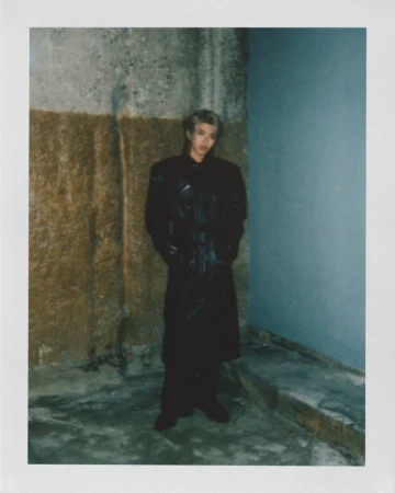 Adult male in black coat poses in dim interior, walls with mixed textures, and a dark ambiance highlighted by a polaroid frame.