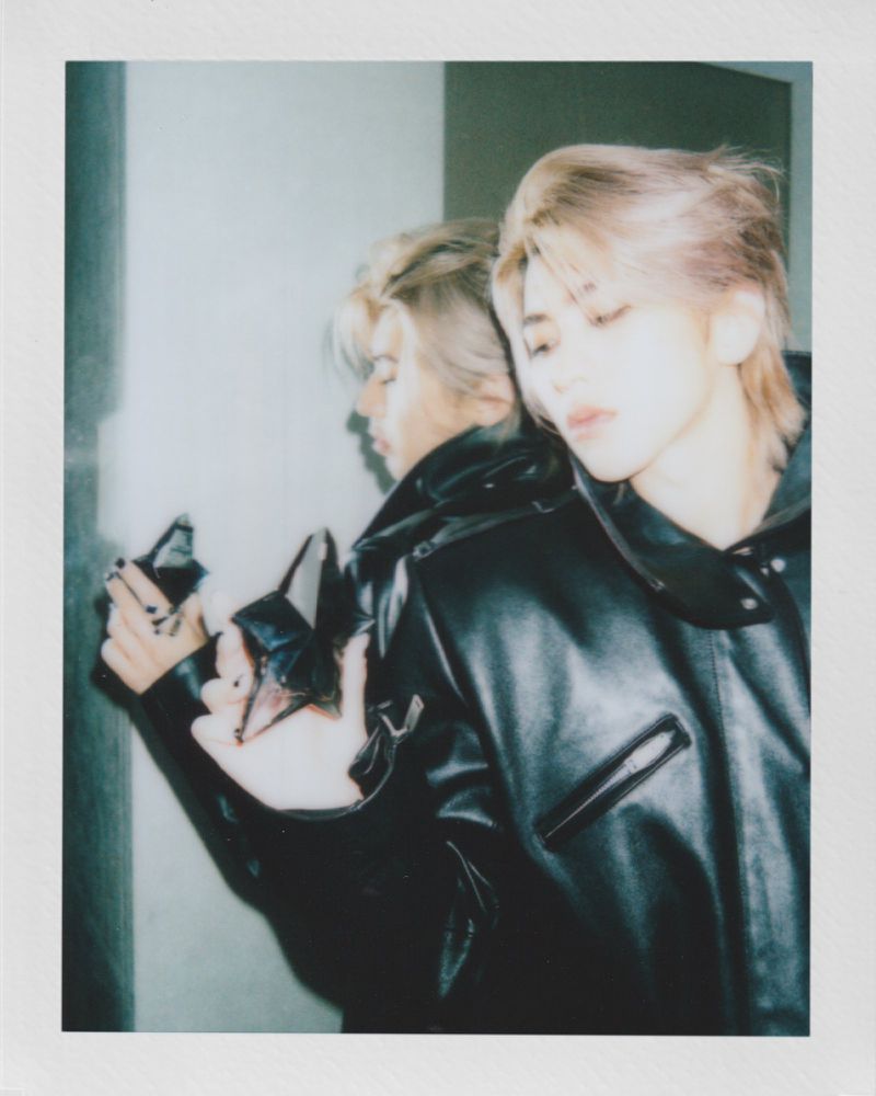 Portrait of a young adult with blonde hair in a black leather jacket, reflected in a mirror, against a neutral grey indoor wall.