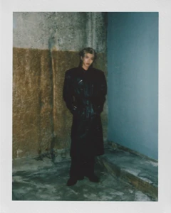 Polaroid portrait of Kun standing against a concrete wall wearing an oversized black leather trench coat.