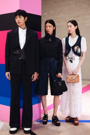 Three Asian fashion models in runway looks pose against a color-blocked background. One wears a suit, another dark separates, the third a lace dress.