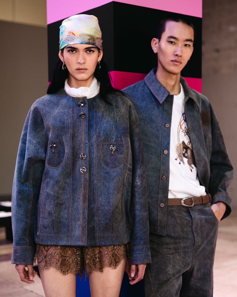Two models pose indoors wearing matching grey denim jackets and pants, with a colorful block pattern in the background.