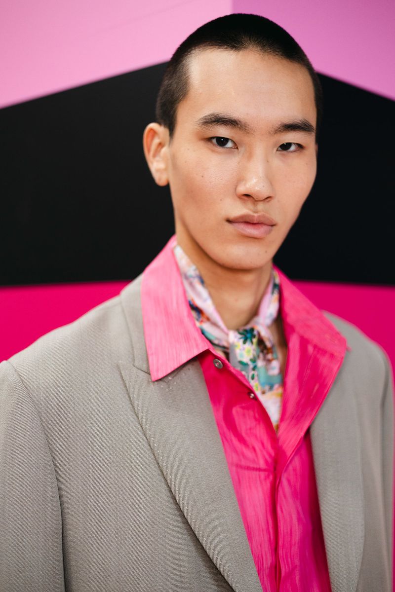 An Asian man in a gray blazer and bright pink shirt stands against a geometric backdrop, showcasing fashion-forward style and modern grooming.