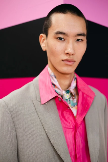 An Asian man in a gray blazer and bright pink shirt stands against a geometric backdrop, showcasing fashion-forward style and modern grooming.