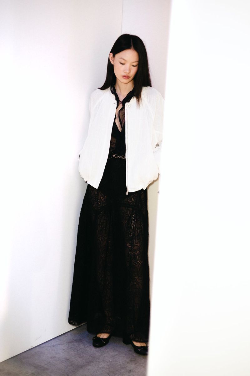 Full-length shot of a slender Asian woman in a black lace dress and white jacket, leaning against a white corner in soft, diffused light.