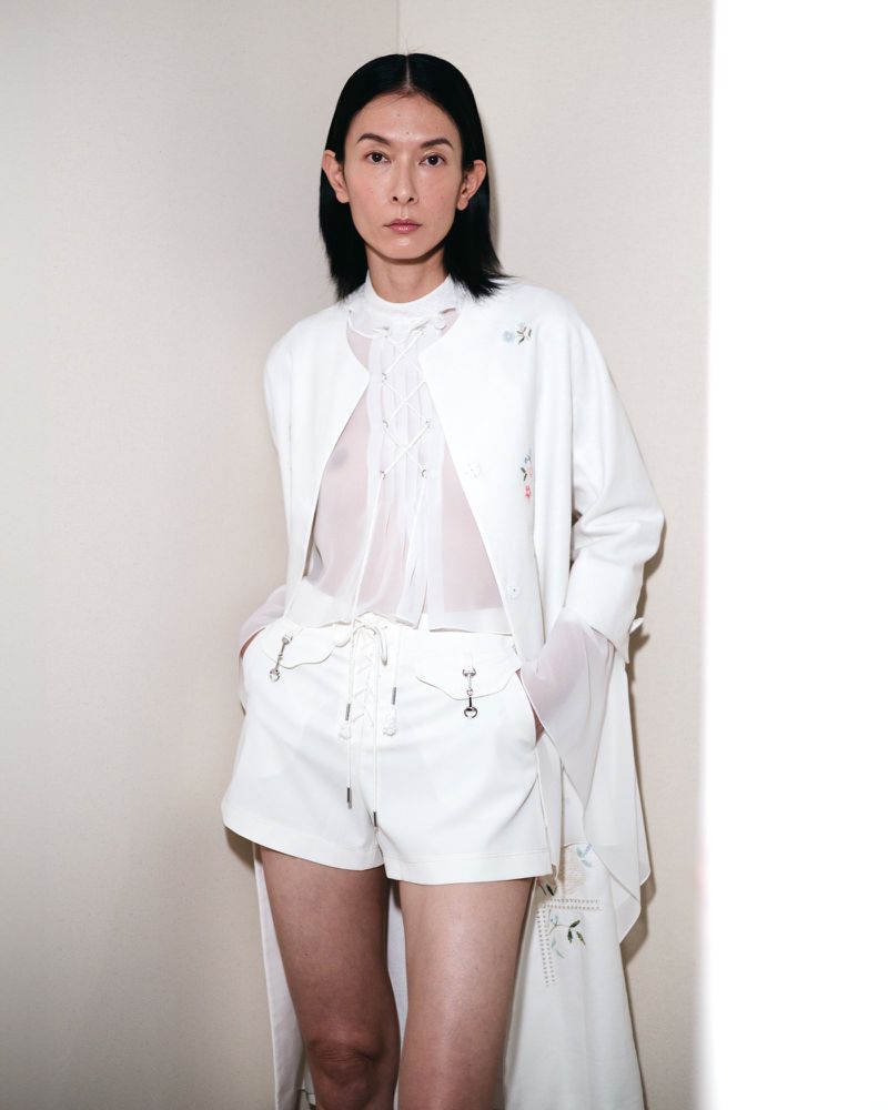 Person poses in a white ensemble consisting of an embroidered jacket, a sheer blouse, and tailored shorts against a neutral wall.