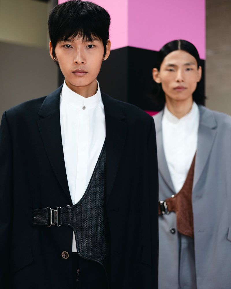 Two androgynous models pose indoors wearing blazers and vests with buckled straps. One model wears a black blazer and one wears a grey blazer.