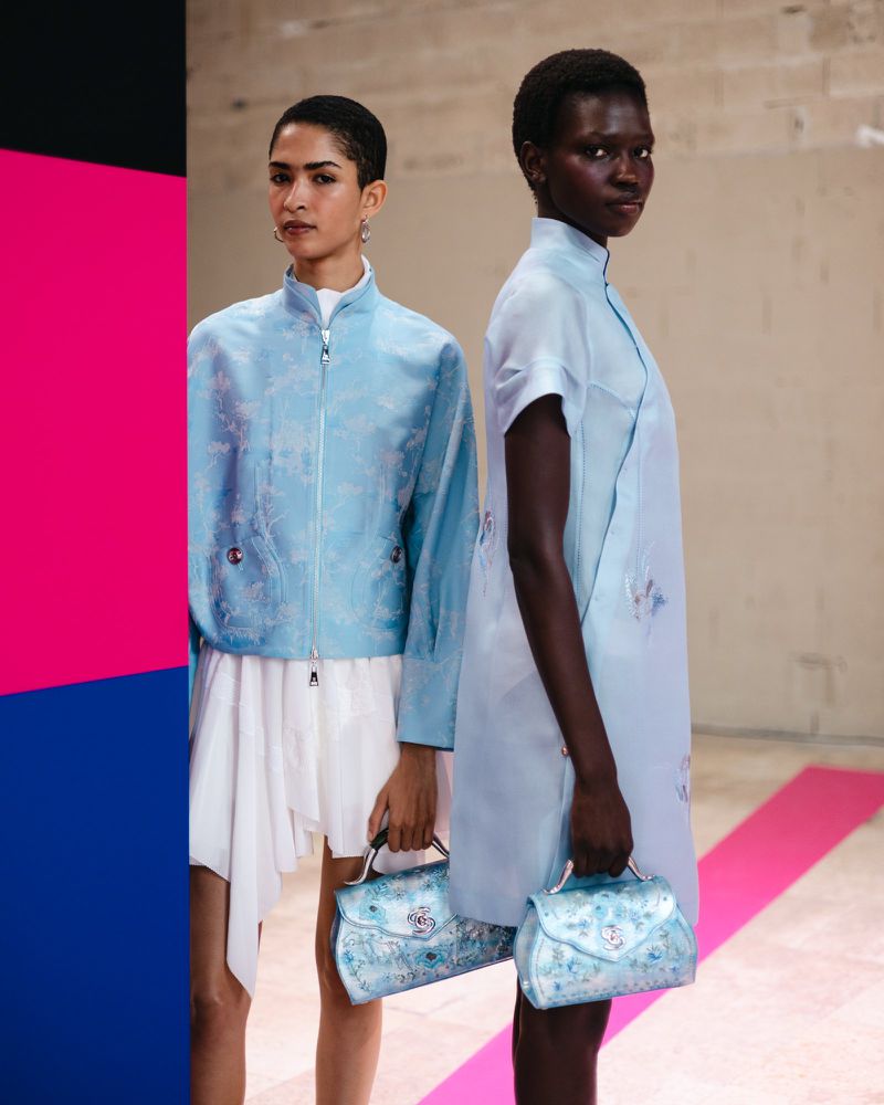 Two diverse fashion models pose indoors with light blue patterned jackets and dresses, holding matching blue floral handbags, editorial.