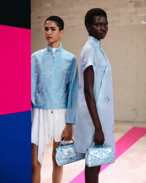 Two diverse fashion models pose indoors with light blue patterned jackets and dresses, holding matching blue floral handbags, editorial.