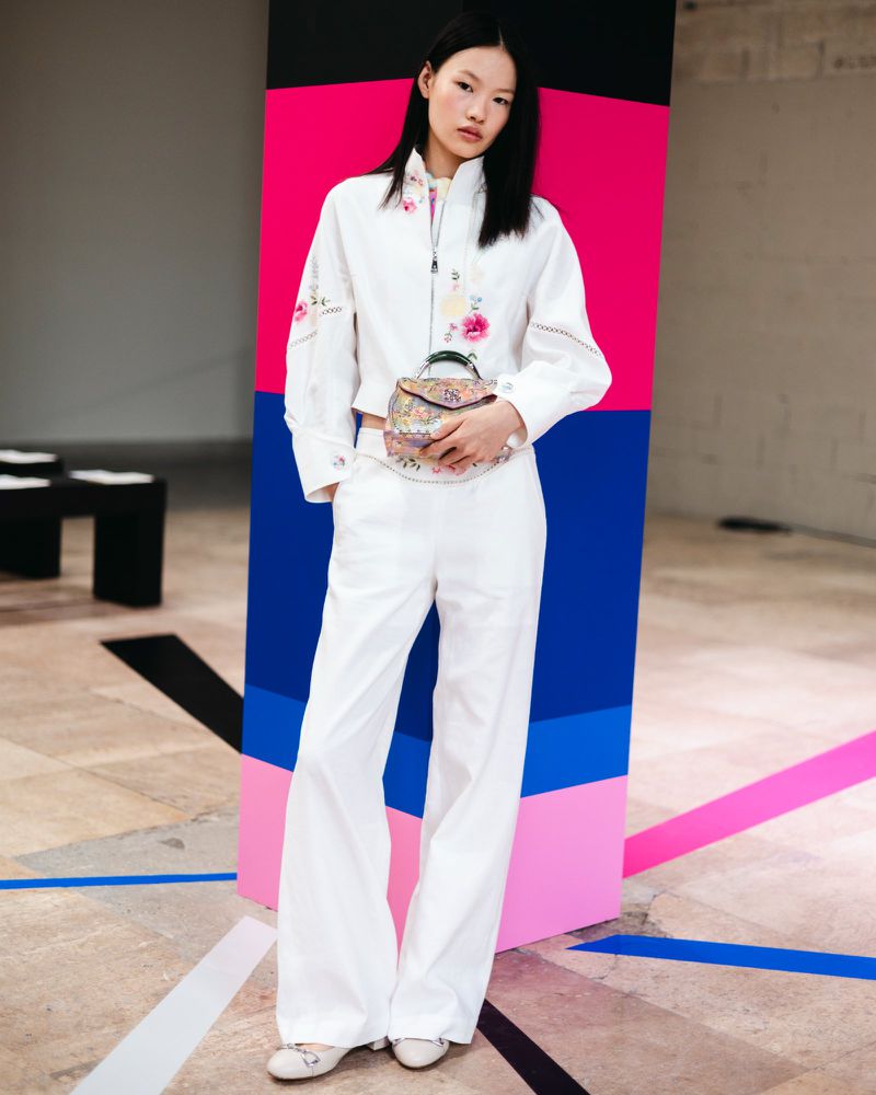 Young woman in white suit with floral purse and jacket stands against a geometric pink, blue, and black background indoors.