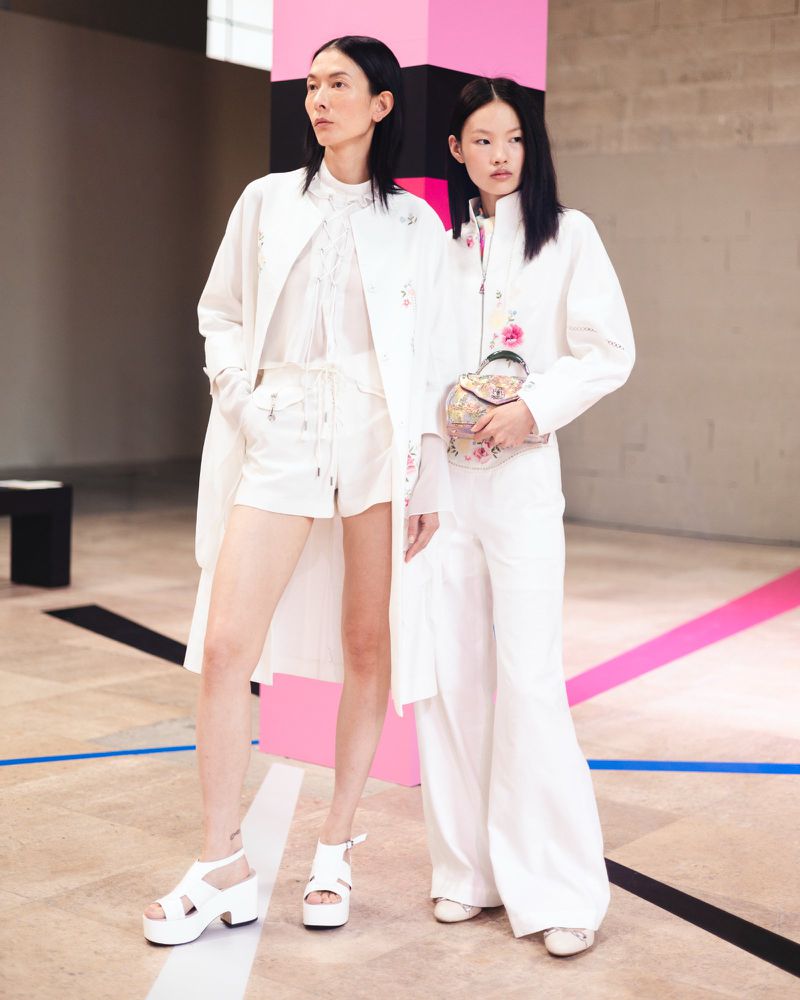 Two Asian fashion models in white outfits pose in an indoor studio with geometric lines and colorful accents, displaying a modern, stylish look.