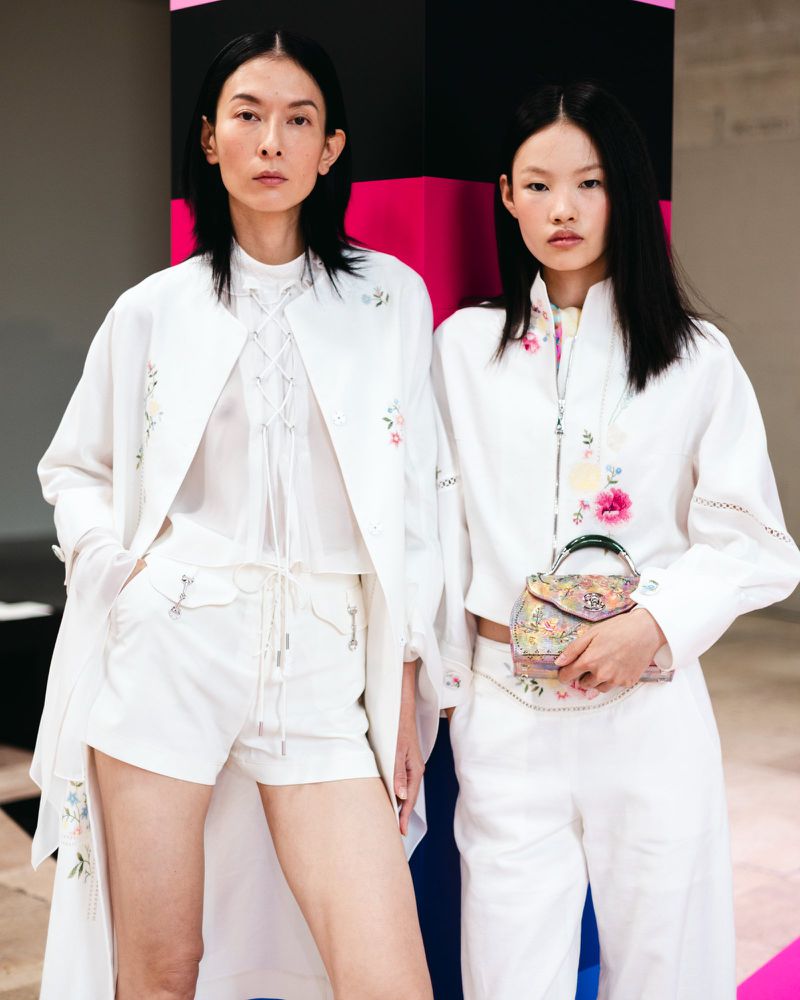 Two female fashion models in all-white floral-embroidered outfits pose against a vibrant pink, black and blue background in a studio setting.