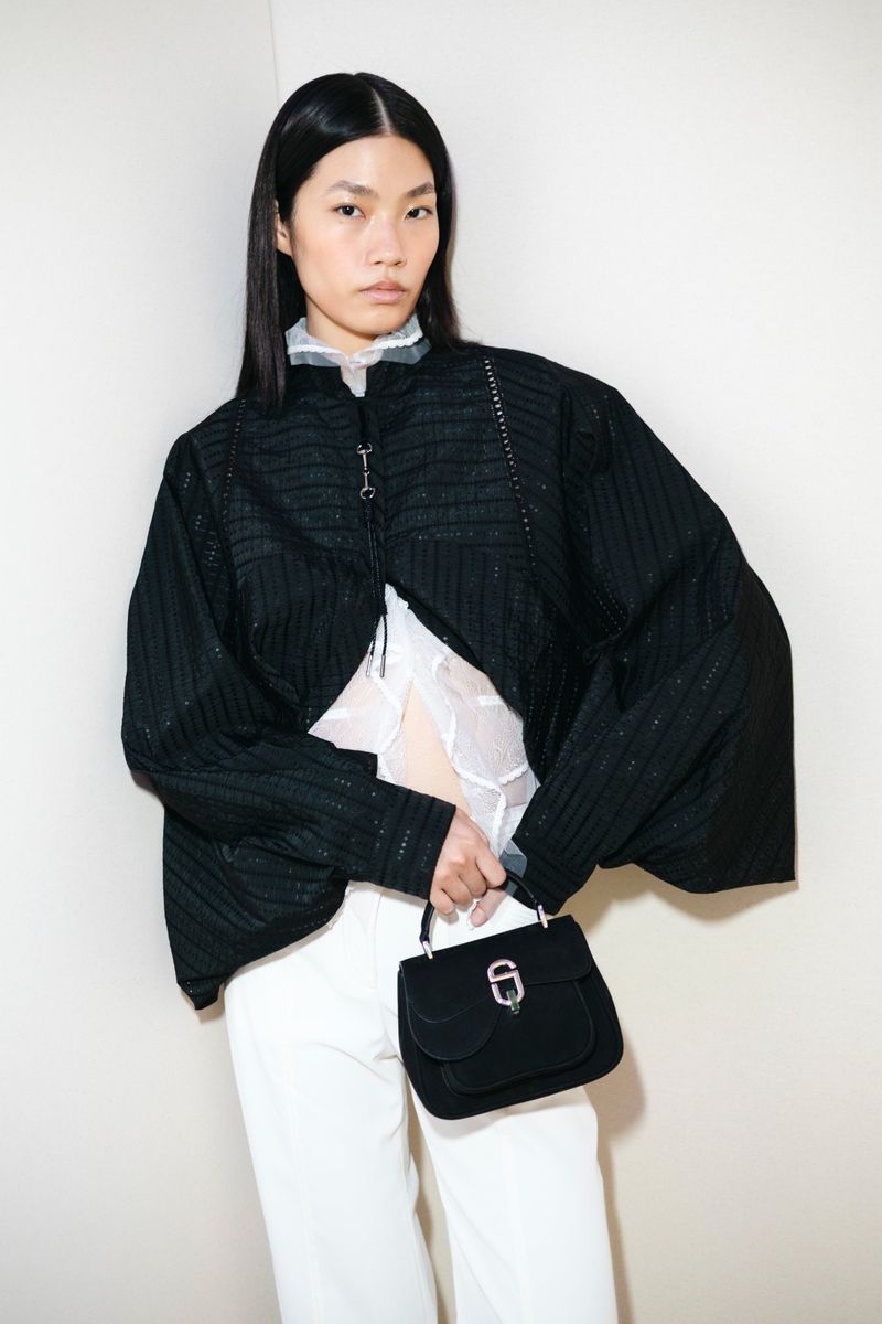 Model wearing a black structured jacket and white trousers holding a black handbag against a plain white wall
