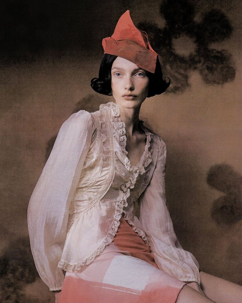 Woman with dark hair poses in a lace-trimmed shirt and unique orange hat against a textured brown background with shadow details.