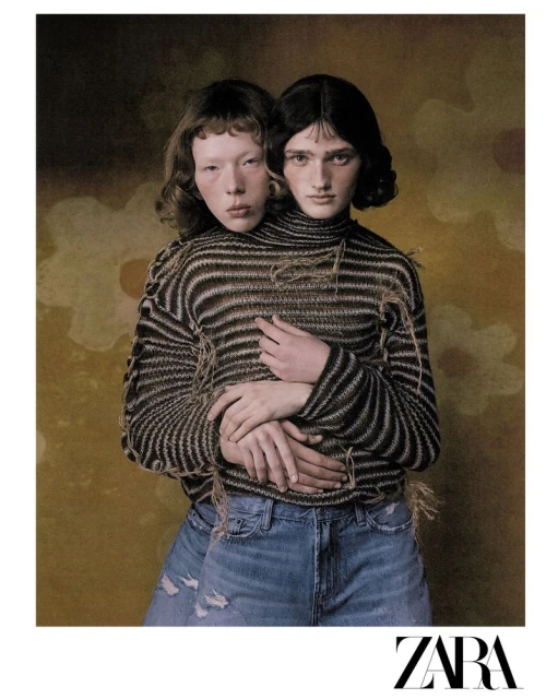 Two models embrace while wearing a striped sweater and blue jeans against a painterly floral backdrop, showcasing fashion and editorial style.