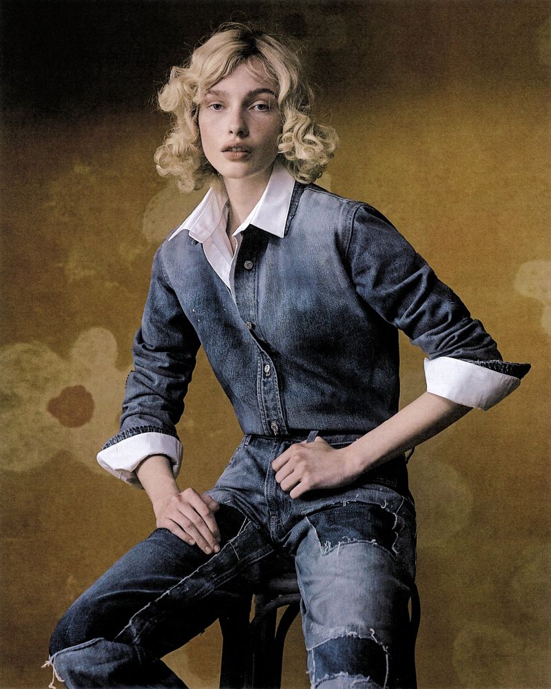 A young adult female poses in a denim outfit with a white collared shirt while sitting on a chair against a mottled brown background.