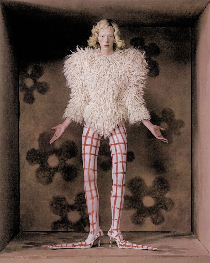 Woman in unusual outfit posing in a studio. She is wearing a furry sweater and shoes/leggings with a red plaid pattern.