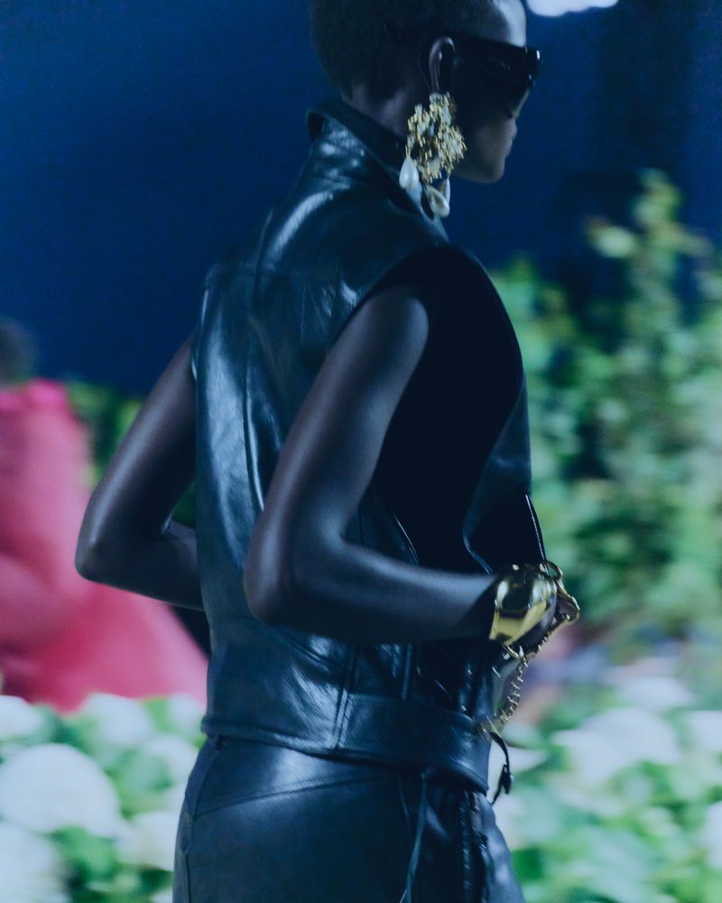 Runway fashion featuring a Black model in a sleeveless leather top, dark shades, and gold jewelry, set against a blurred floral backdrop.