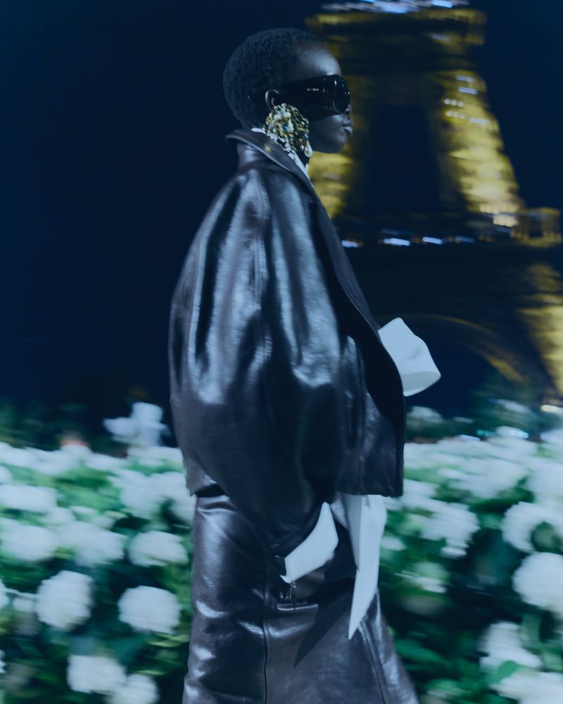 Fashion runway show featuring an African female model in a black leather skirt and jacket, large earrings, and Paris Eiffel Tower backdrop.
