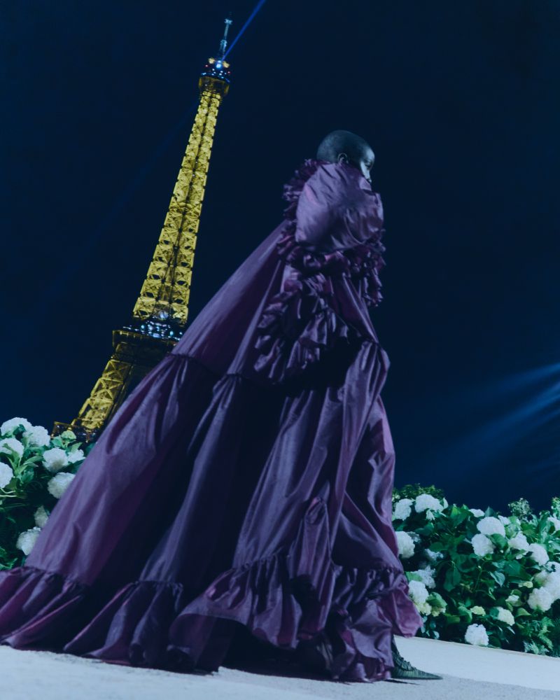 Fashion photography of a woman in a ruffled gown on a runway, with the Eiffel Tower illuminated in the background against a night sky.