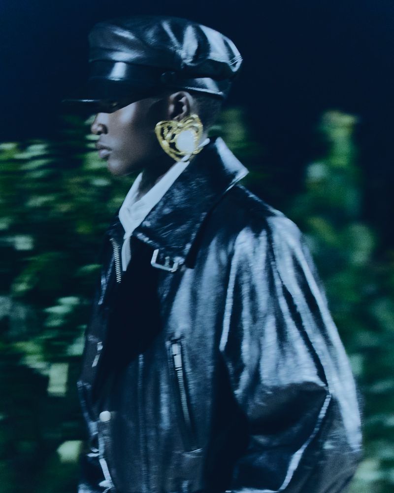 Portrait of Black woman in black patent-leather jacket and hat, wearing heart-shaped gold earrings with white pearl, blurry green background.