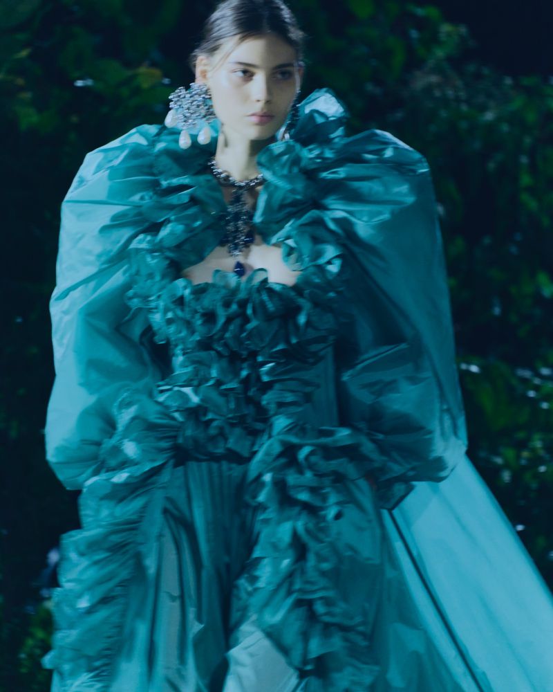 A woman with fair skin and light brown hair wears a ruffled, teal-colored gown, statement earrings and necklace, posing in a garden setting.