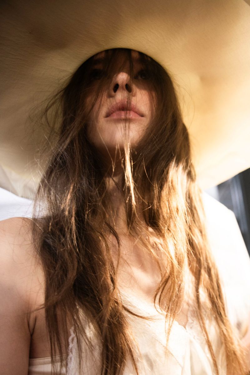 Portrait of a woman with long brown hair and a large, light-colored hat. She is looking directly at the camera in an indoor setting.