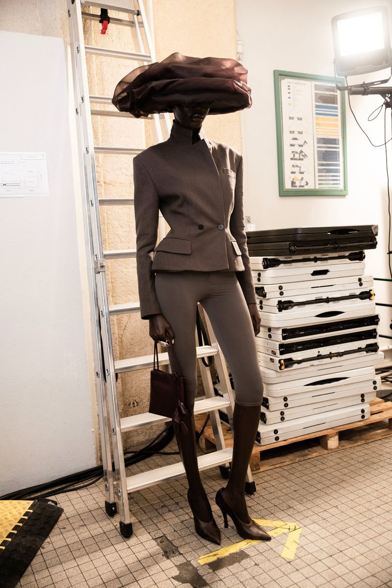 Fashion shot of a Black model in a structured grey suit and large hat holding a purse, posing in front of a ladder and studio equipment.