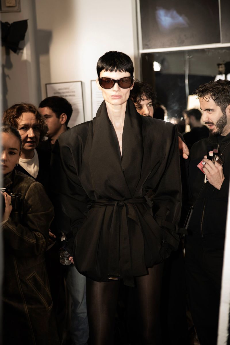 A woman with short dark hair and sunglasses poses in a black wrap jacket with a belt, surrounded by blurred figures in an indoor setting.