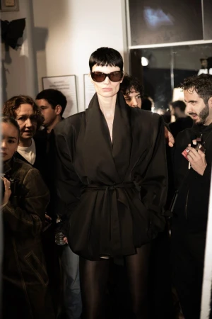 A woman with short dark hair and sunglasses poses in a black wrap jacket with a belt, surrounded by blurred figures in an indoor setting.