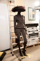 Model wearing a structured tailored blazer, cropped leggings, pointed heels, and an oversized dark puff hat posing backstage.