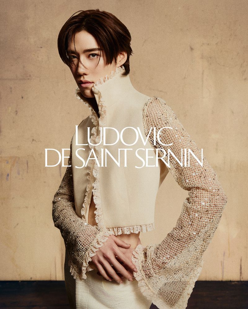 A person with brown hair poses wearing a white outfit with lace sleeves. 'Ludovic de Saint Sernin' text overlay against tan background.