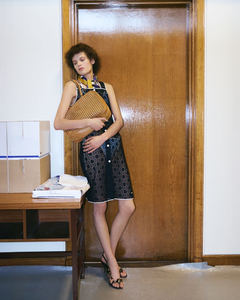Woman in a black dress with a woven tote bag, standing by a wooden door and boxes. Fashion editorial photography with natural lighting.