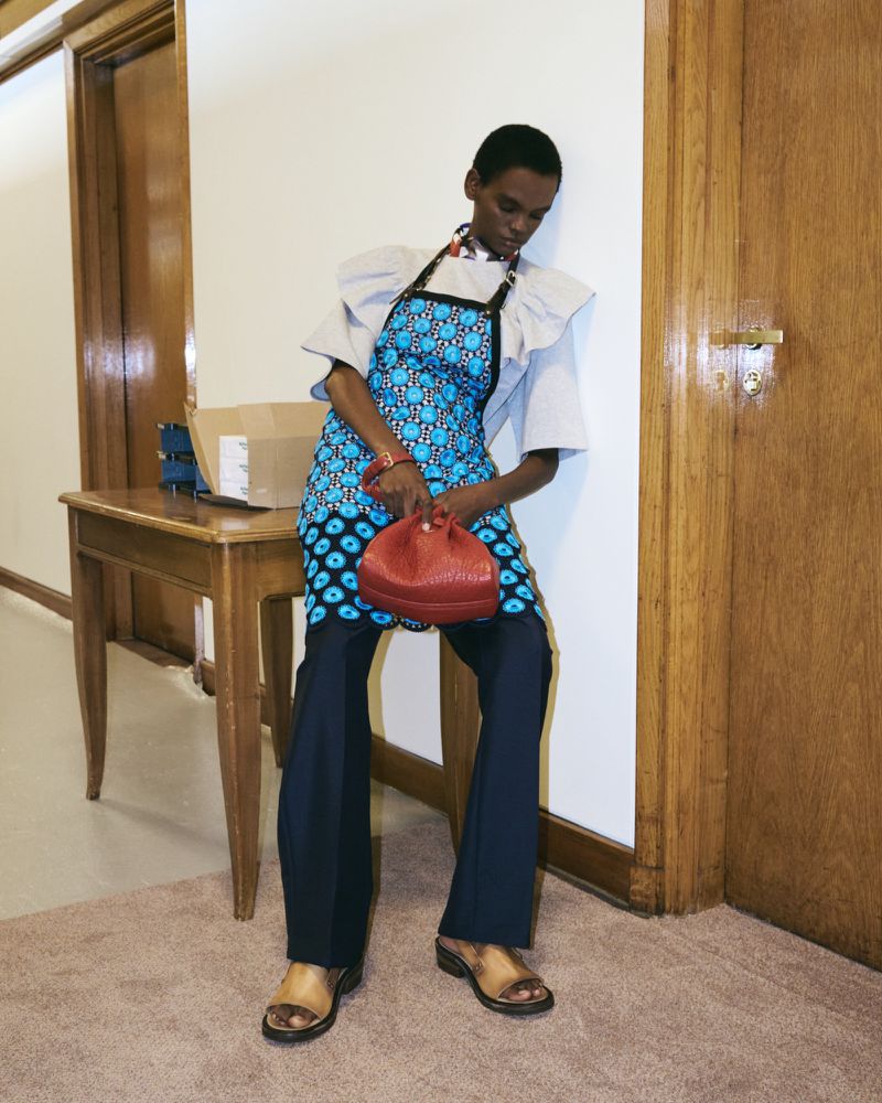 Female model wearing an apron-style dress with a blue pattern, holding a red purse, and posing in an indoor setting against a wall.