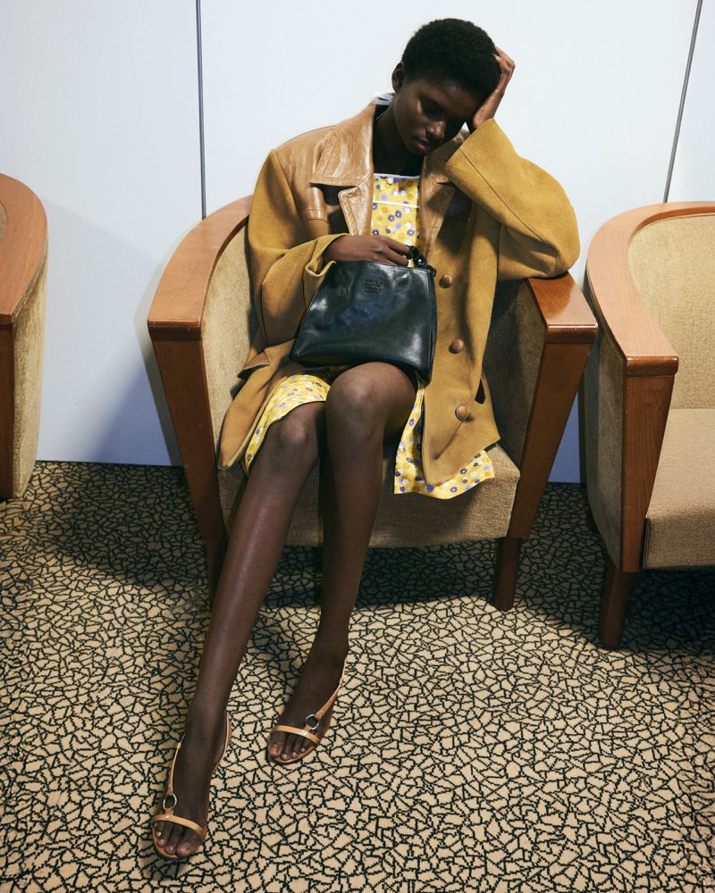 A young, stylish woman in a mustard-colored coat sits in a chair holding a black purse with legs crossed in patterned interior setting.