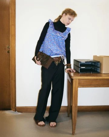 Woman in vintage fashion posing in indoor setting with a desk, wearing a blue floral print top, black pants, sandals and holding a purse.