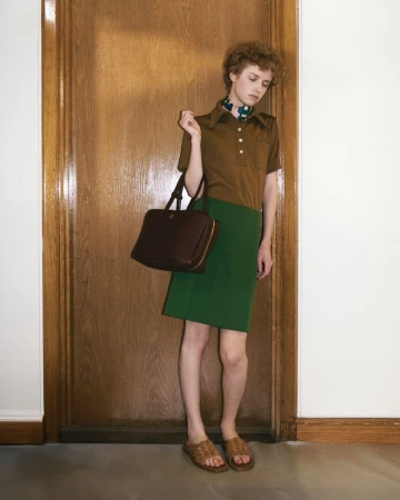 A person stands in front of a closed wooden door wearing a brown shirt, green skirt, sandals, and holding a brown bag.