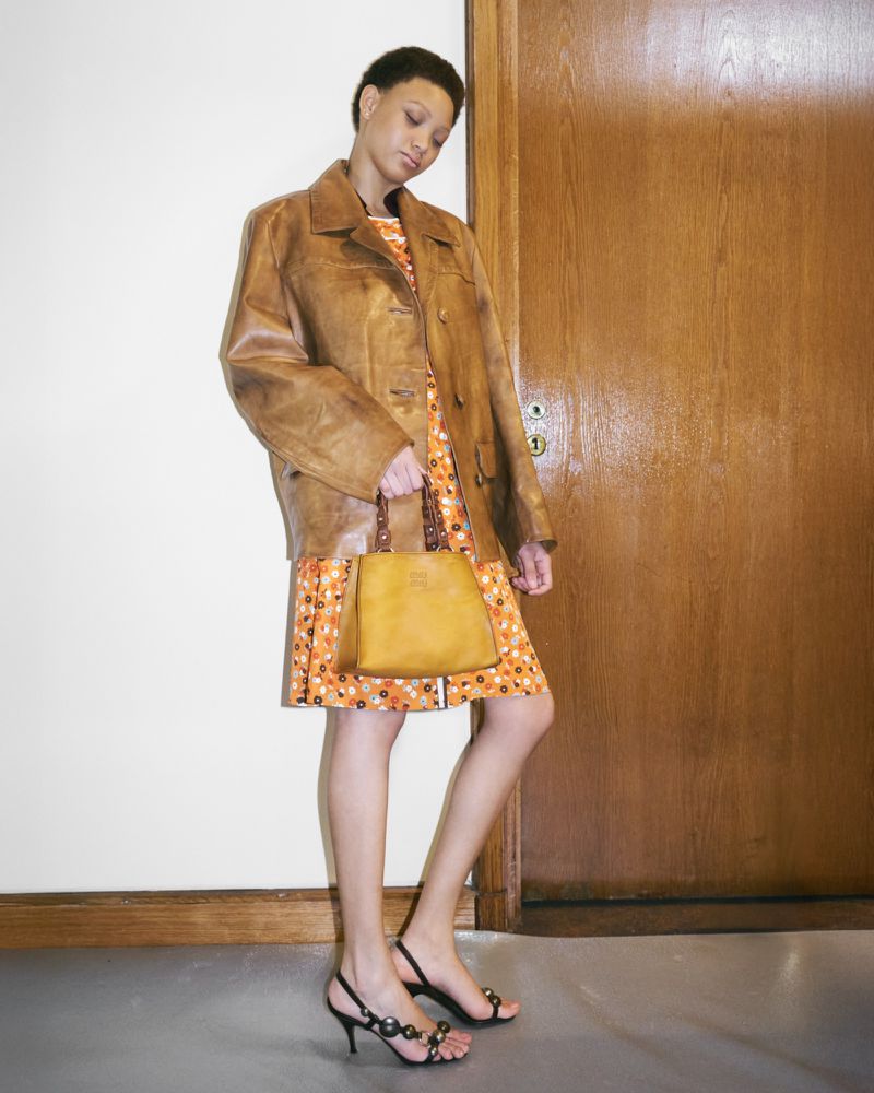 Model wearing a brown leather jacket and patterned dress holding a handbag indoors.