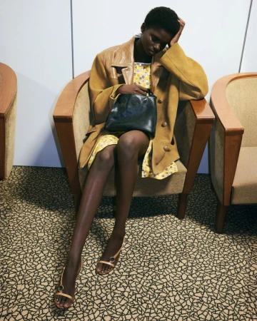 Model seated in an armchair wearing an oversized mustard coat, floral slip dress, and strappy sandals holding a black leather handbag.