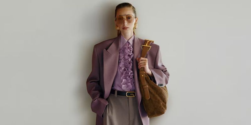 A fashionable woman in a purple blazer, ruffled blouse, and sunglasses poses against a neutral wall, holding a textured shoulder bag.