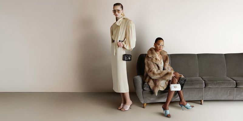Two women in a studio setting, one seated in a faux fur coat and the other standing in a draped shirt and skirt, both holding purses.