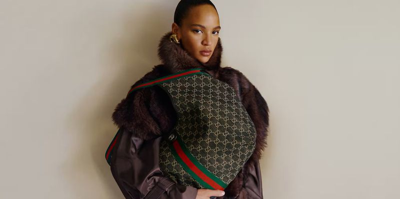 Woman wearing a patterned Gucci bag with red and green stripes, and a brown fur collared jacket poses in a studio with soft lighting.