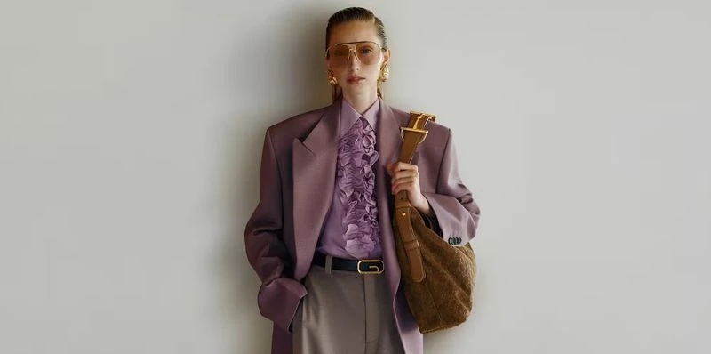 Model wearing tinted aviator sunglasses, a plum oversized blazer, and a brown suede shoulder bag against a neutral grey wall.