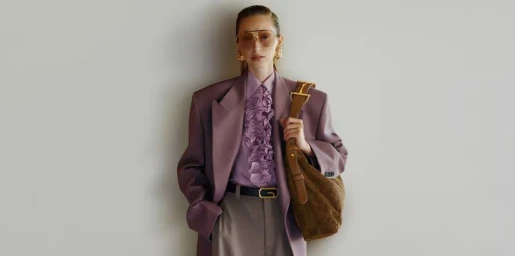 Model wearing tinted aviator sunglasses, a plum oversized blazer, and a brown suede shoulder bag against a neutral grey wall.
