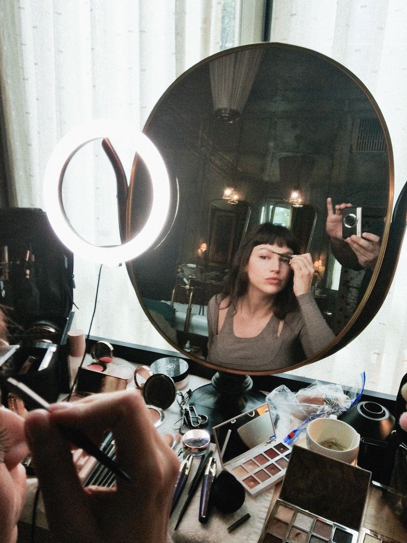 Adult female applying makeup in front of a round mirror with a ring light on a vanity with cosmetic products and brushes in an indoor setting.